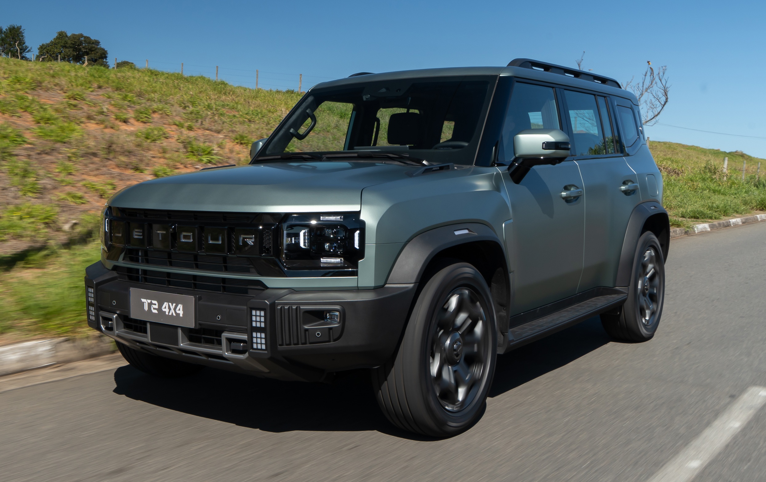 SUV Jetour T2 4x4 cinza-esverdeado em movimento, visto de frente e lateral, em uma estrada asfaltada com gramado e c&eacute;u azul ao fundo
