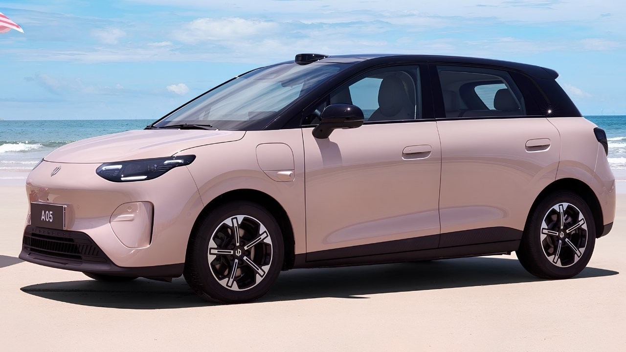 Carro elétrico rosa claro com teto preto, modelo A05, estacionado na areia da praia, com o mar azul e o céu claro ao fundo