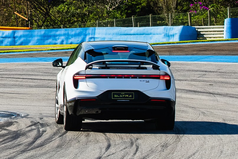 Traseira de um carro branco Lotus Eletre em uma pista de corrida, com faixas azuis e amarelas, e vegeta&ccedil;&atilde;o ao fundo