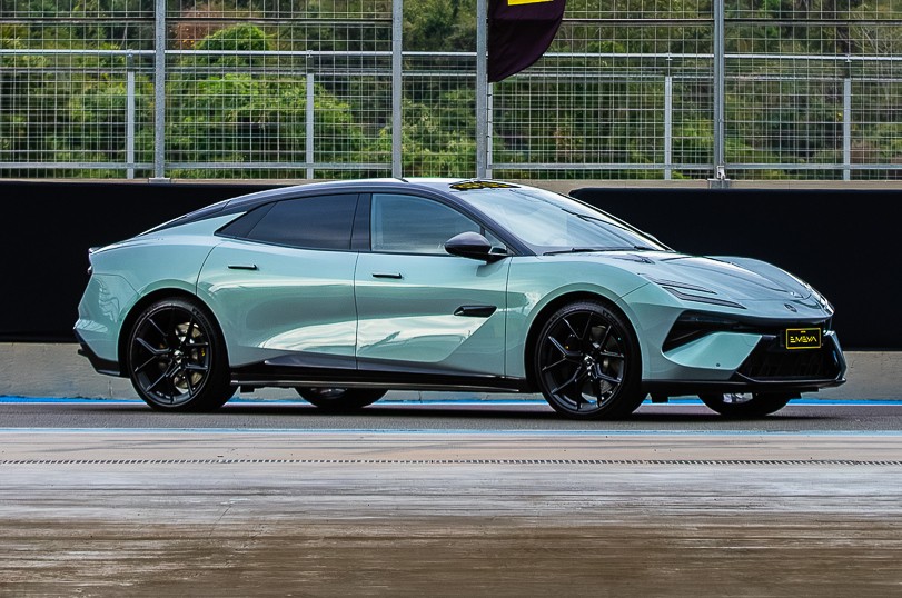 Carro esportivo el&eacute;trico Lotus Emeya azul-claro met&aacute;lico com teto preto, rodas pretas e detalhes pretos, parado em uma pista de corrida com grade e &aacute;rvores ao fundo