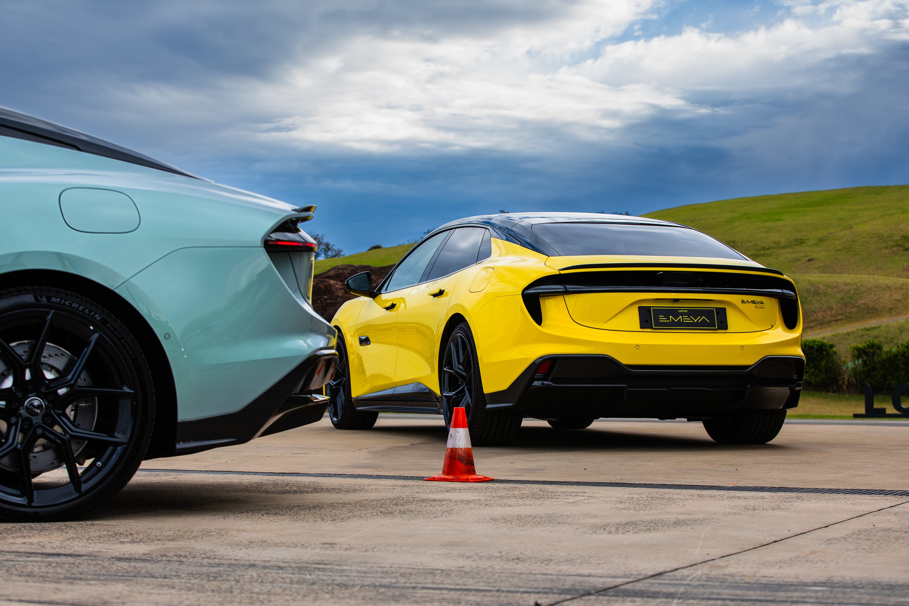 Dois carros esportivos modernos, um verde-&aacute;gua &agrave; esquerda e um amarelo &agrave; direita, estacionados em uma pista de asfalto. O carro amarelo tem a placa EMEVA e um cone de tr&acirc;nsito laranja &agrave; frente. Ao fundo, colinas verdes e um c&eacute;u nublado com nuvens escuras