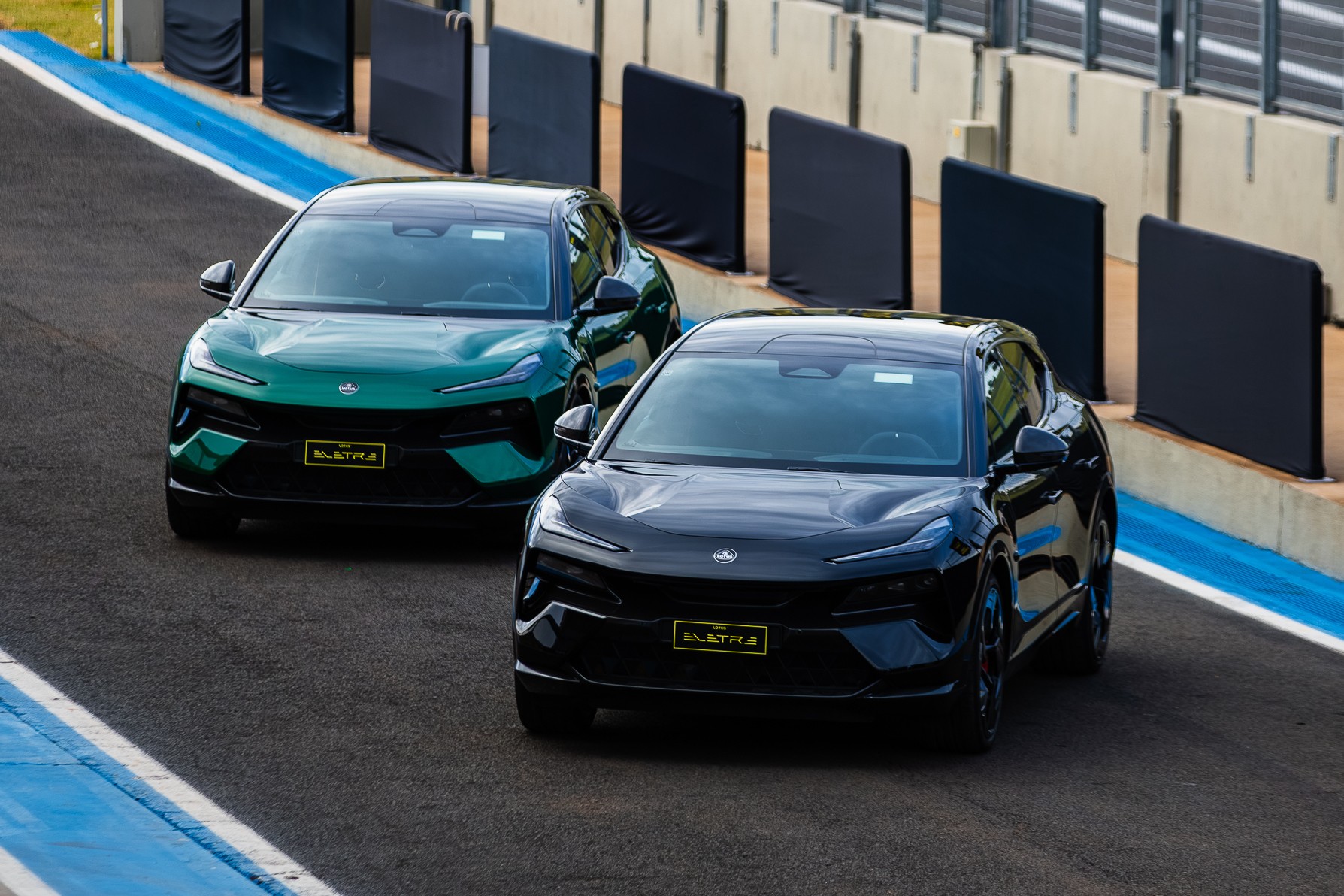 Dois carros esportivos Lotus Eletre, um verde escuro e outro preto, em uma pista de corrida com barreiras de prote&ccedil;&atilde;o ao fundo
