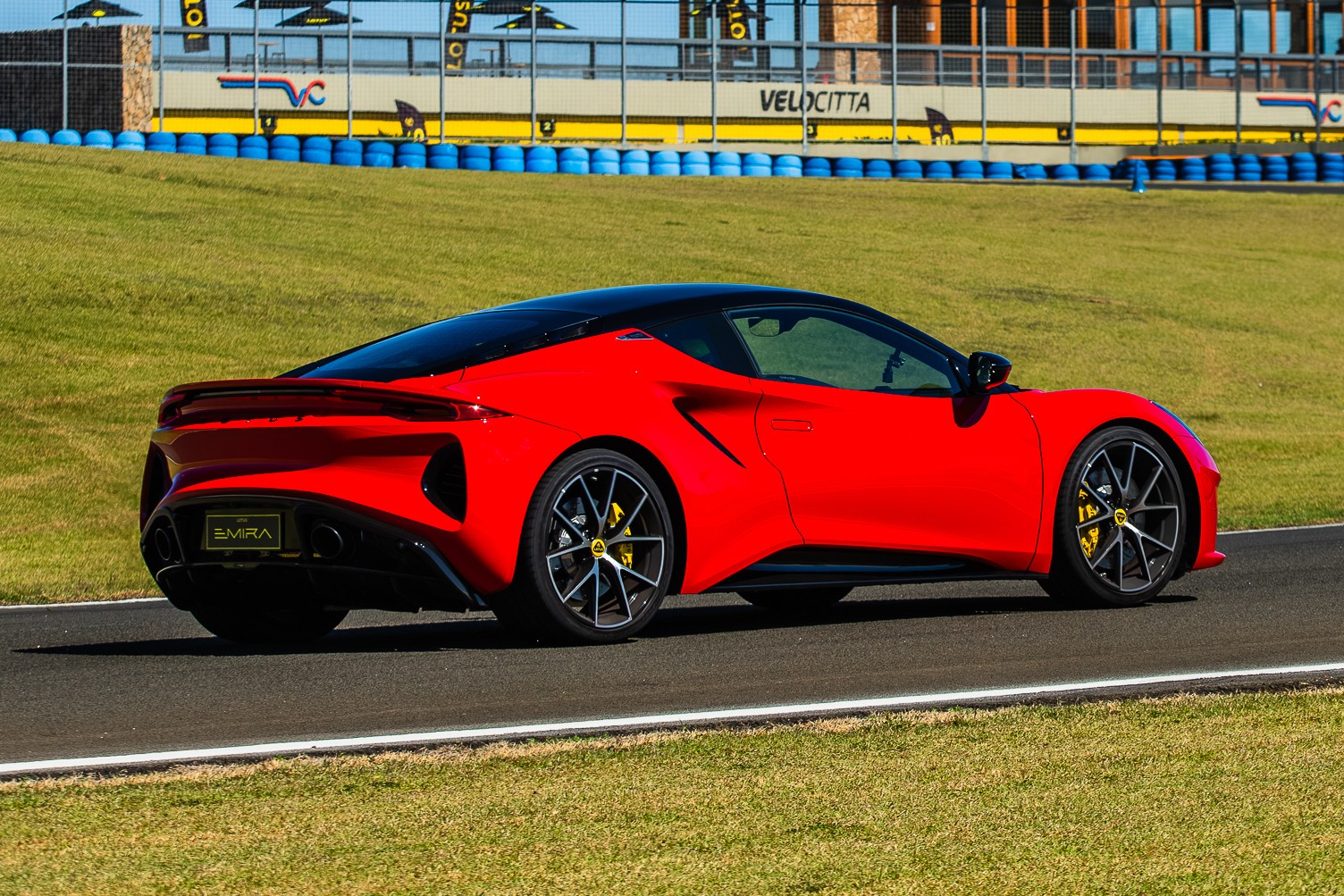 Carro esportivo vermelho com teto preto, modelo Lotus Emira, parado em uma pista de corrida. Ao fundo, uma barreira de pneus azuis e uma constru&ccedil;&atilde;o com a palavra VELOCITTA