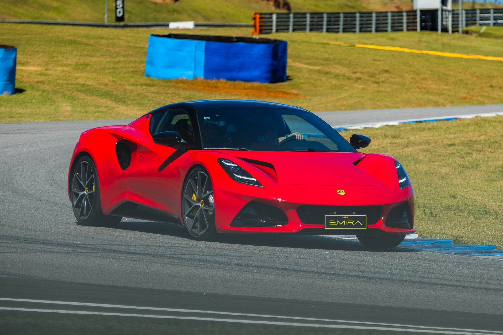 Carro esportivo Lotus Emira vermelho com teto preto, placa EMIRA e rodas escuras com pin&ccedil;as amarelas, em uma pista de corrida cinza com faixas brancas e azuis, gramado verde ao fundo
