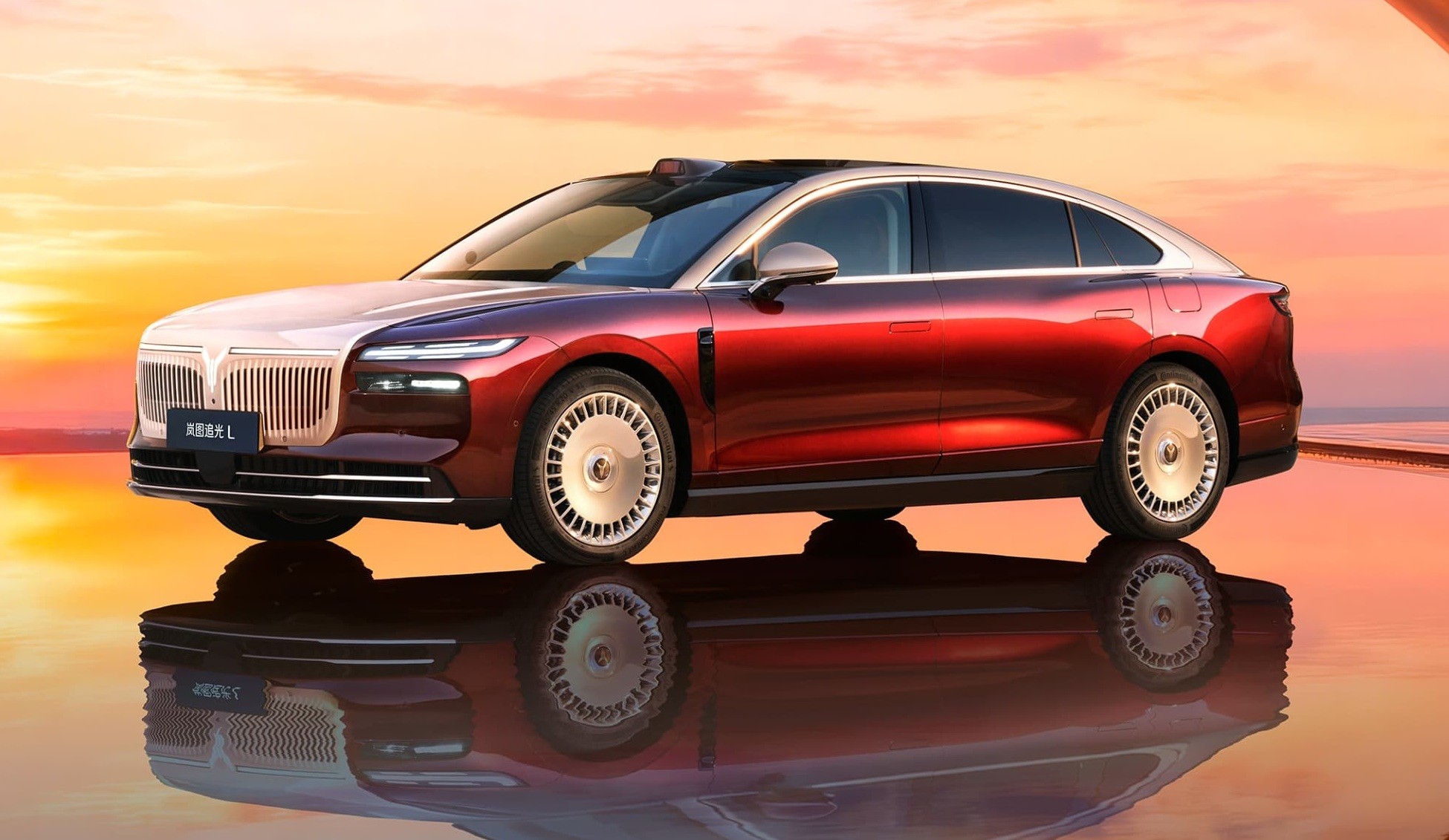 Carro de luxo vermelho e branco com teto preto, parado sobre superf&iacute;cie espelhada que reflete sua imagem, contra um c&eacute;u alaranjado de p&ocirc;r do sol
