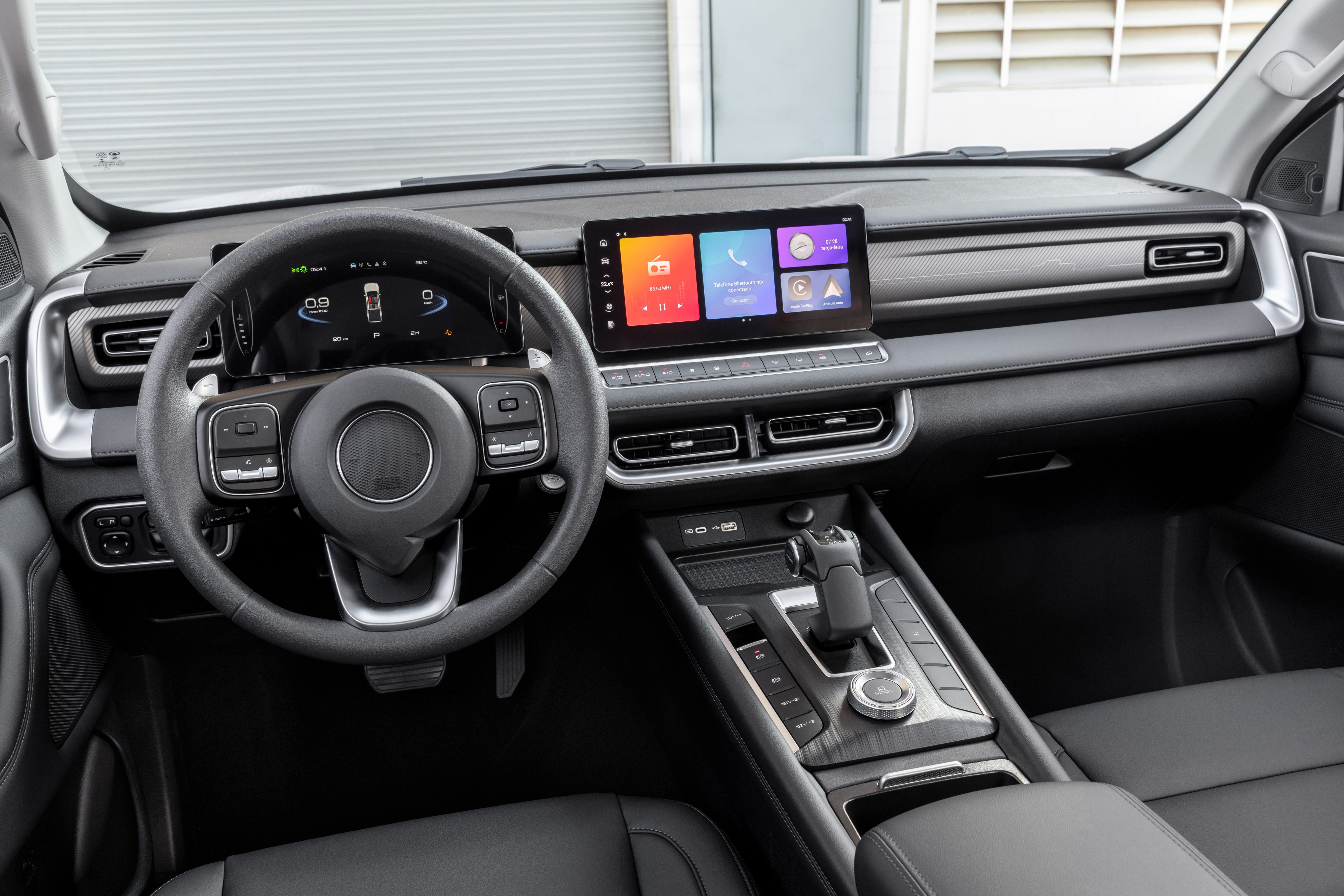 Interior de um carro moderno com painel preto e detalhes prateados. Volante multifuncional, painel digital e tela multim&iacute;dia central com &iacute;cones coloridos. C&acirc;mbio autom&aacute;tico e console central com bot&otilde;es e acabamento em madeira escura. Bancos de couro pretos vis&iacute;veis na parte inferior.