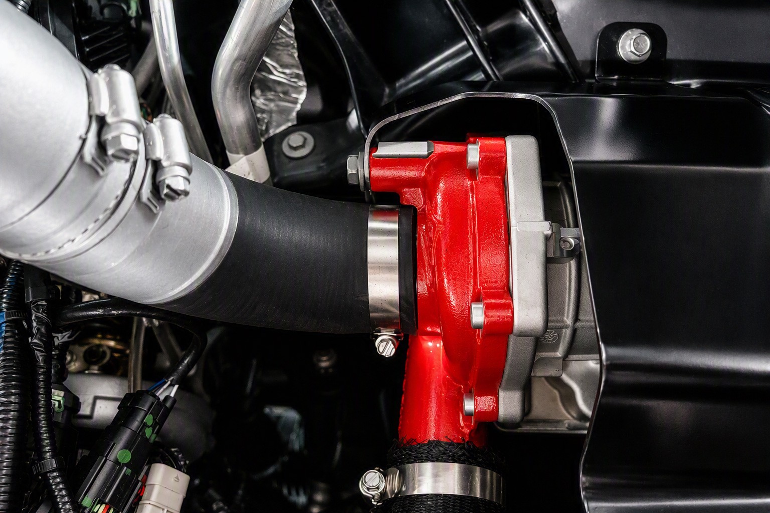 Detalhe do motor de um carro, mostrando um componente vermelho brilhante conectado a uma mangueira preta e um tubo metálico prateado, presos por braçadeiras de metal. Fios e outras peças escuras do motor são visíveis ao fundo