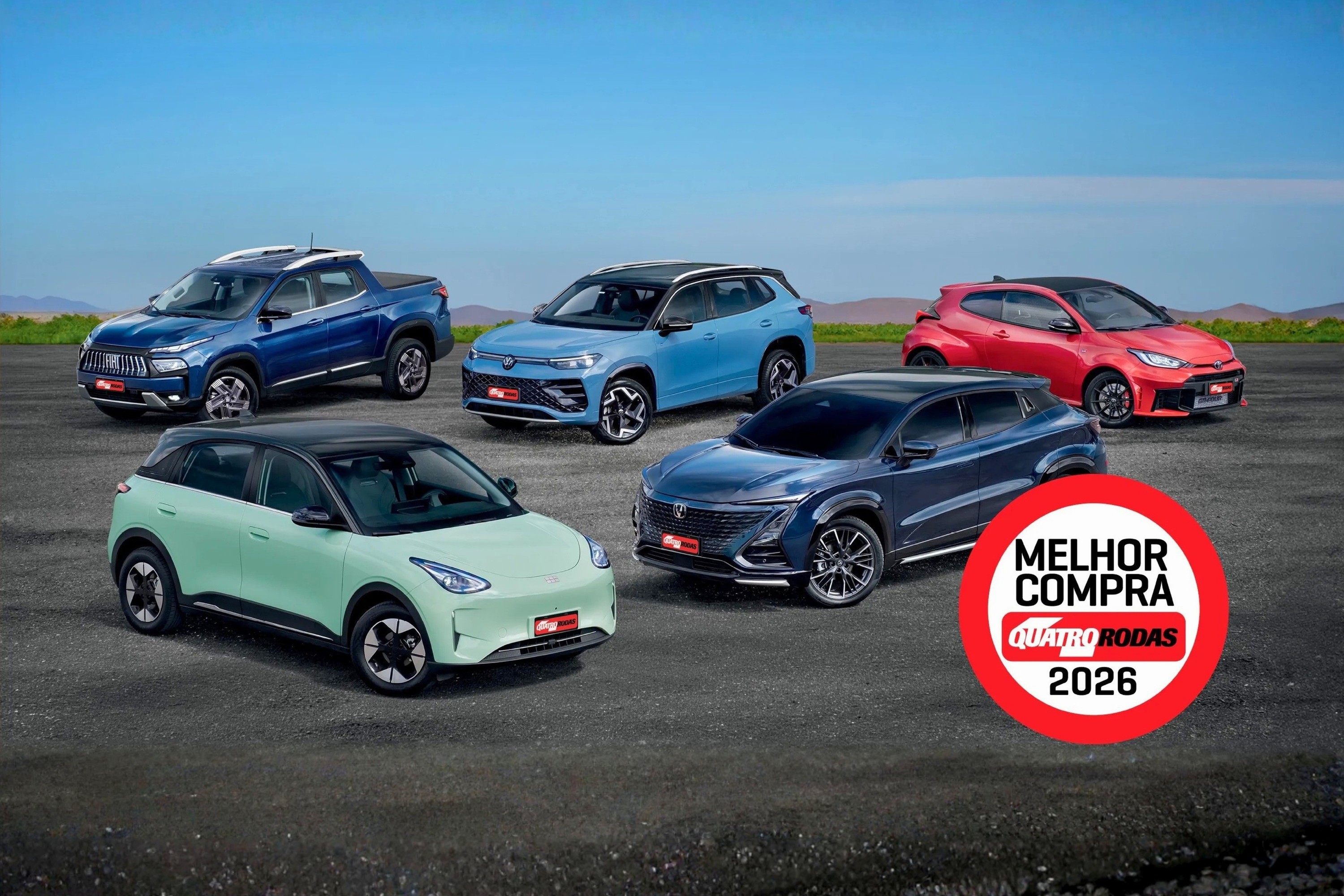 Cinco carros de diferentes modelos e cores, incluindo uma picape azul, um SUV azul-claro, um hatch vermelho, um SUV azul-escuro e um carro verde-claro, estacionados em uma pista de asfalto sob céu azul. No canto inferior direito, um selo vermelho com MELHOR COMPRA QUATRO RODAS 2026 em branco