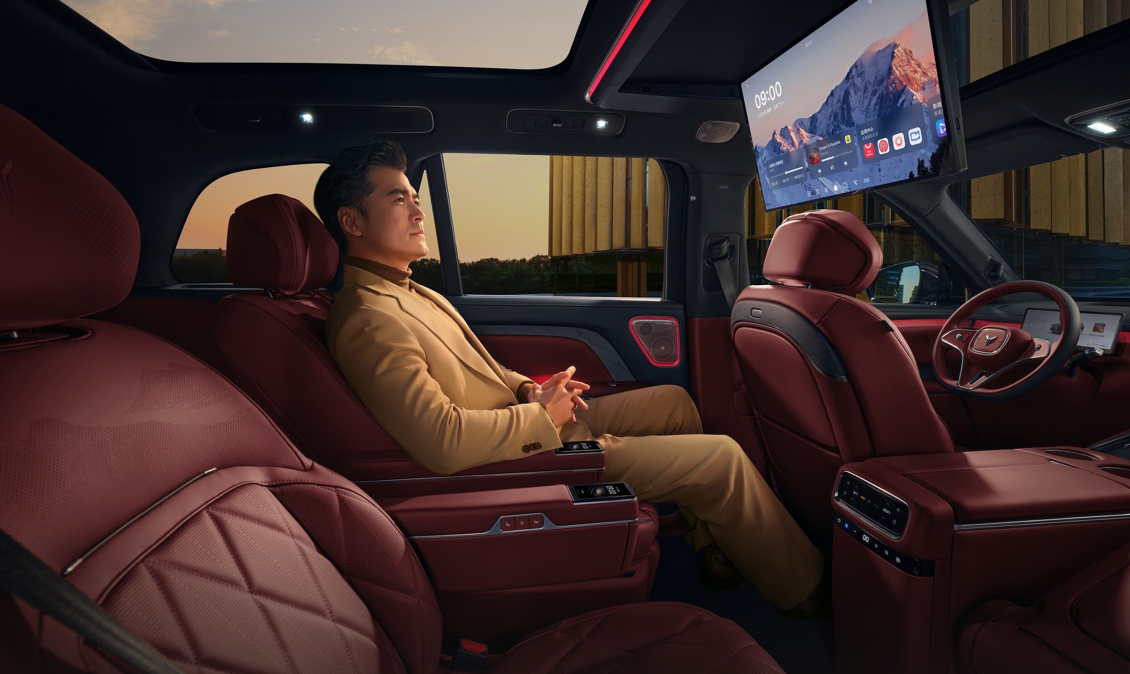 Homem asi&aacute;tico de terno bege relaxa no banco traseiro de carro luxuoso com estofamento vermelho, olhando pela janela. Um monitor grande exibe uma paisagem montanhosa no teto do ve&iacute;culo