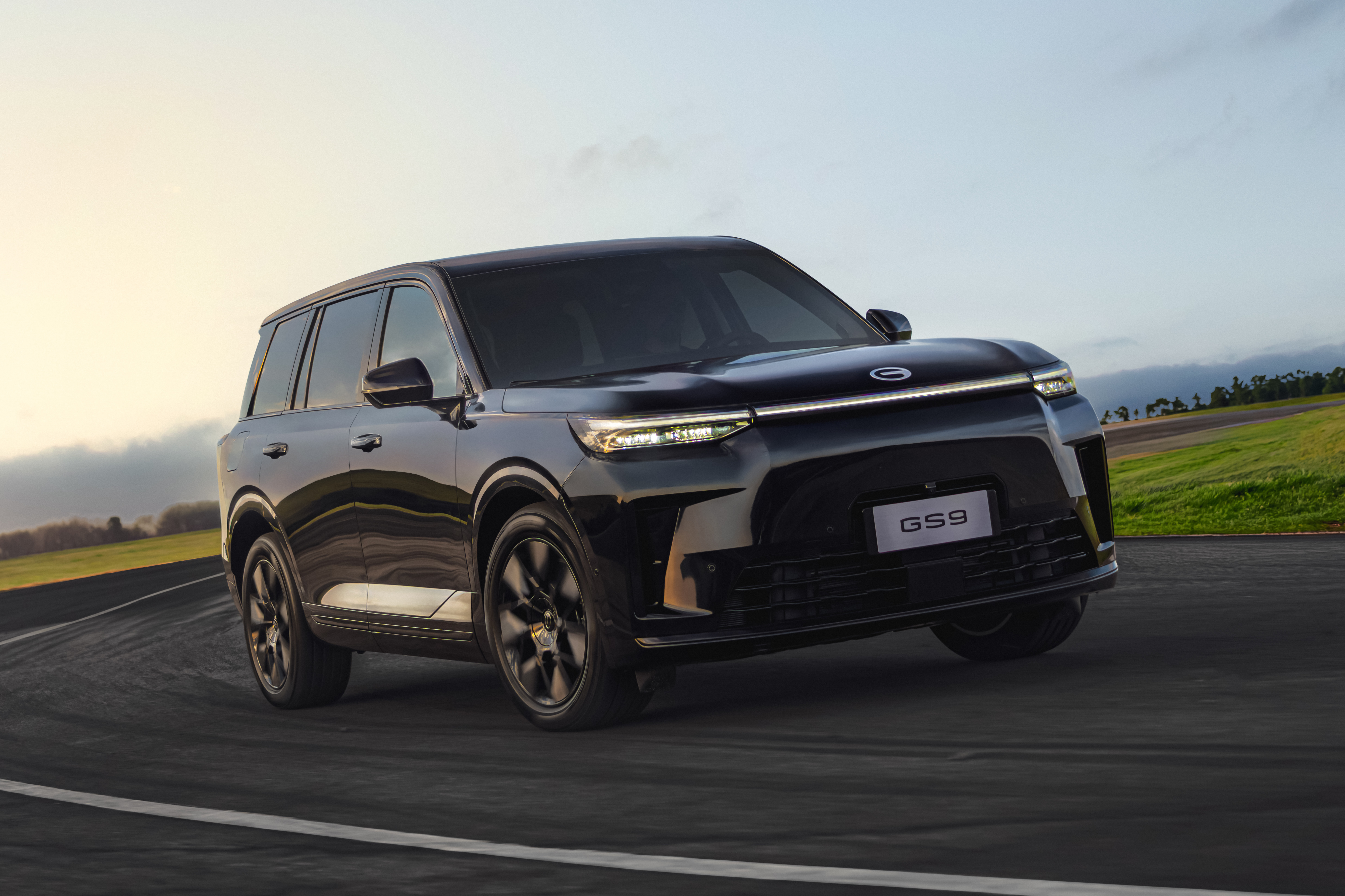 SUV escuro GAC GS9 em movimento numa estrada asfaltada, com campos verdes e c&eacute;u claro ao fundo
