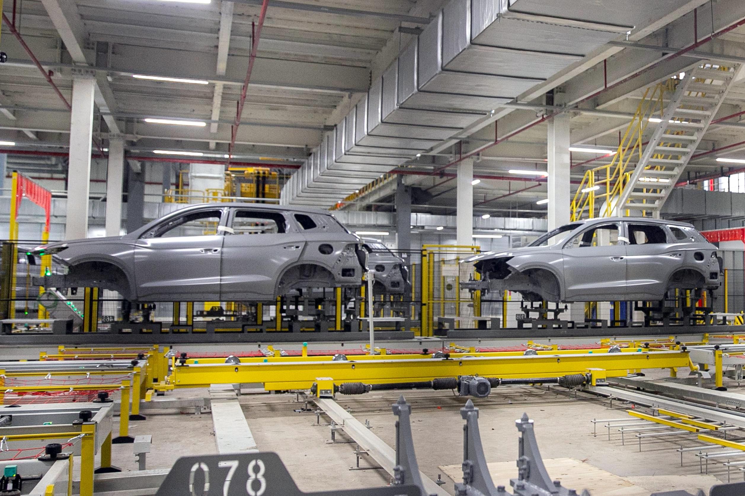 Carroceria cinza de um SUV em linha de montagem industrial, com outra carroceria similar ao fundo. Estruturas metálicas e esteiras transportadoras amarelas preenchem o chão da fábrica, sob um teto alto com dutos e iluminação.